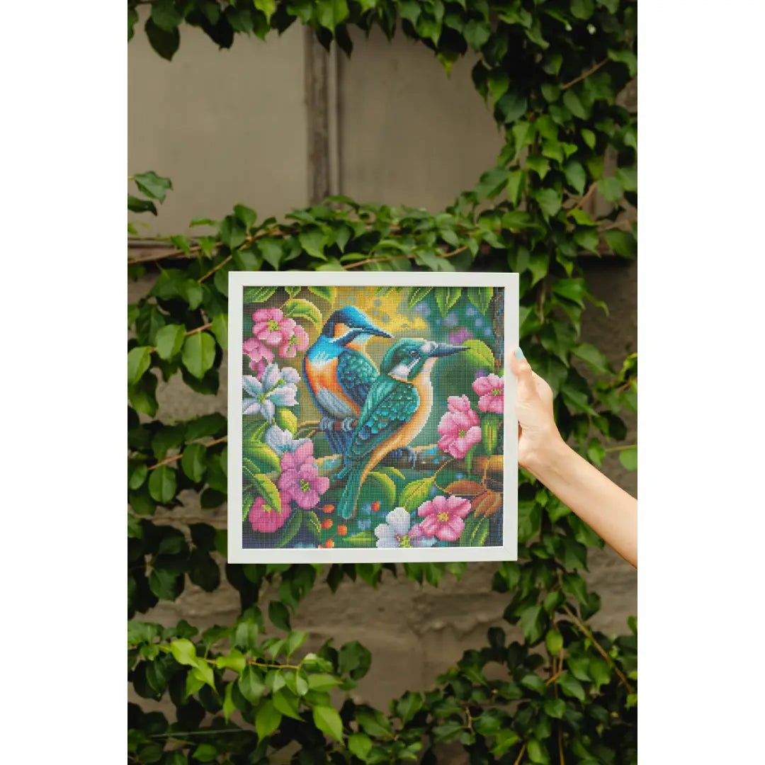 Diamond painting van twee kleurrijke vogels op een tak, omringd door roze en witte bloemen, voor een groene blader achtergrond.