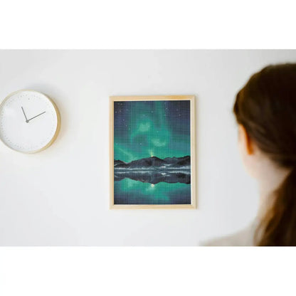 Ingelijste diamond painting van het noorderlicht boven een berglandschap met sterrenhemel en reflectie in een stil meer, hangend aan een witte muur.